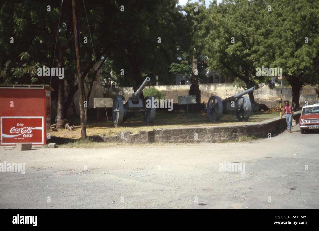 Vernacular photograph taken on a 35mm analog film transparency, believed to depict black car parked near green trees during daytime, 1987. Landmarks/locations detected include Coca-Cola. Major topics/objects detected include Tree, Vehicle, Road, Street and Gray Color. () Stock Photo