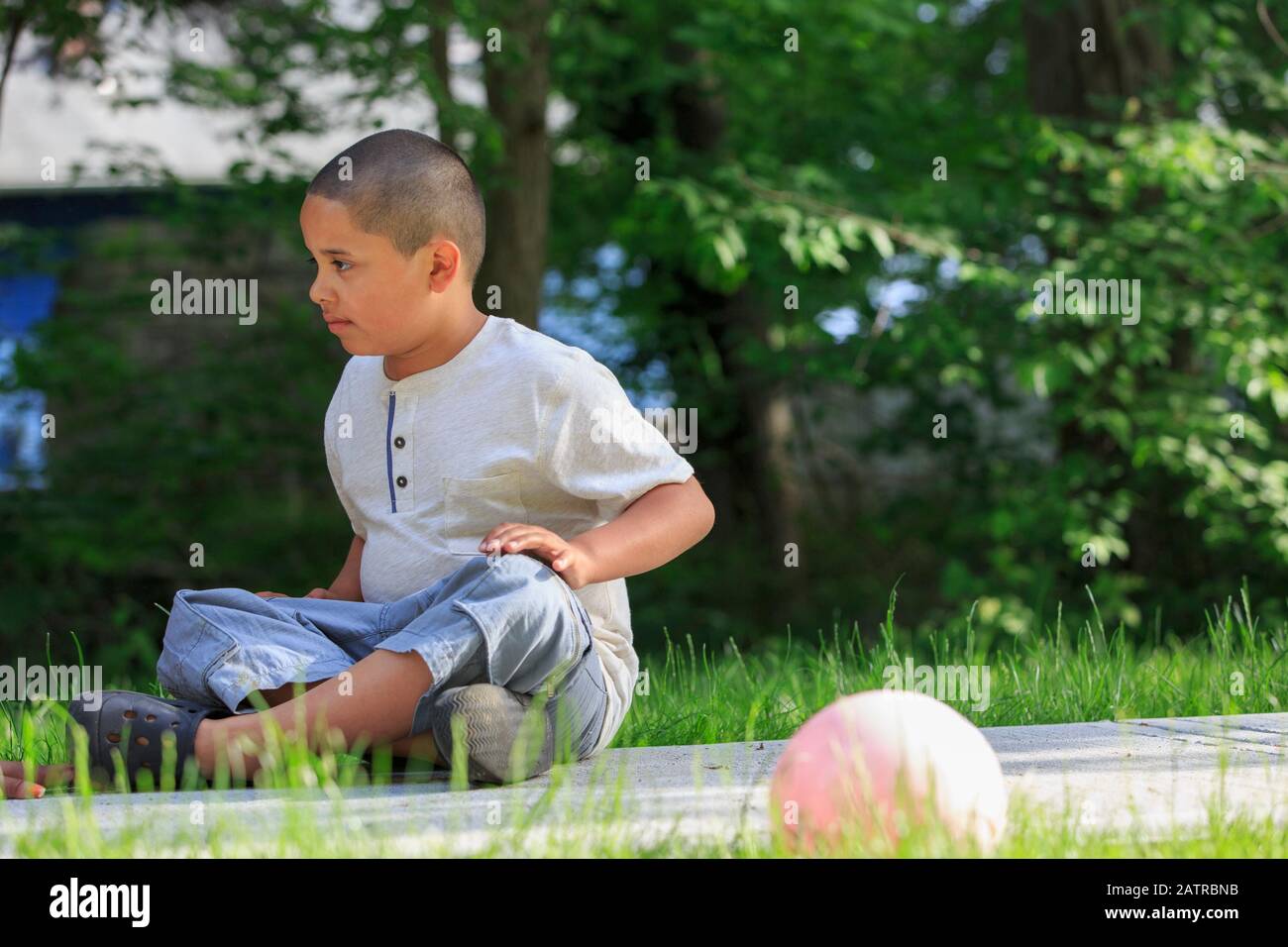 Pre-teen boy outside sitting on the ground with trees in the background Stock Photo