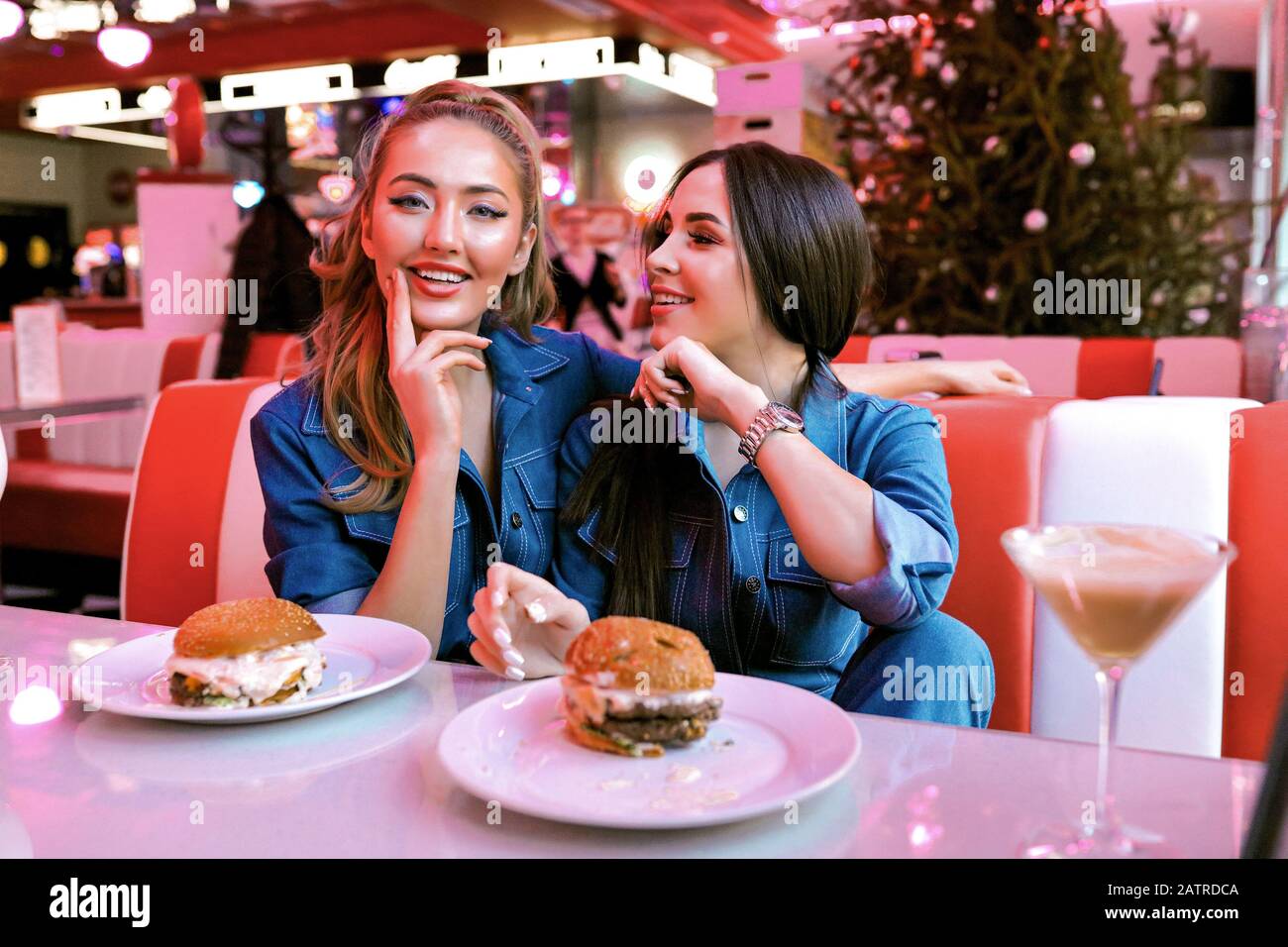 Funny pretty ladies smiling and happy sitting in the restaurant and waiting for their order. Talking and laughing on some jokes.Tasty burgers on plate Stock Photo