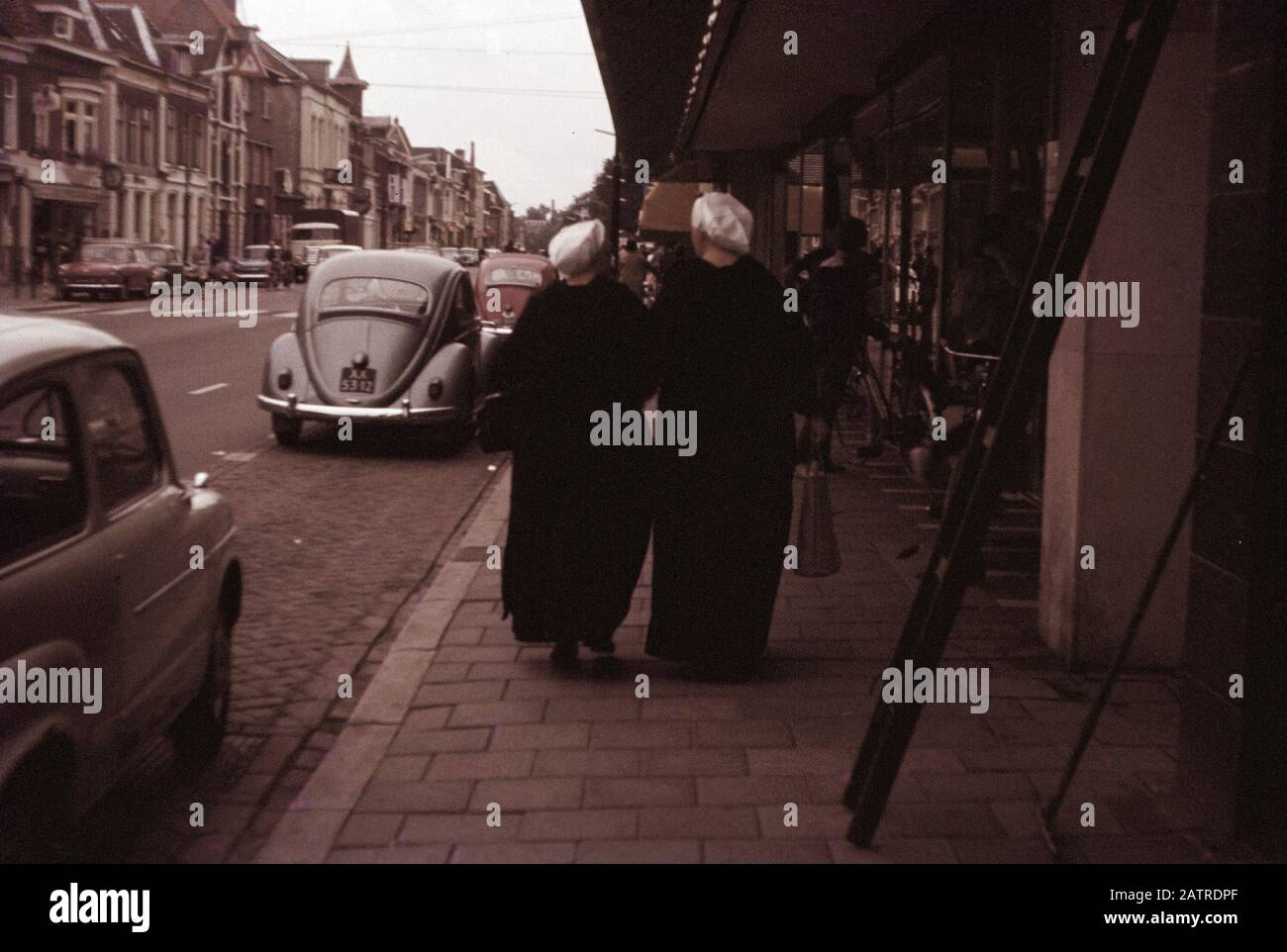 Two mature women wearing long black coats walk down a sidewalk in a city center past cars and houses, 1970. () Stock Photo