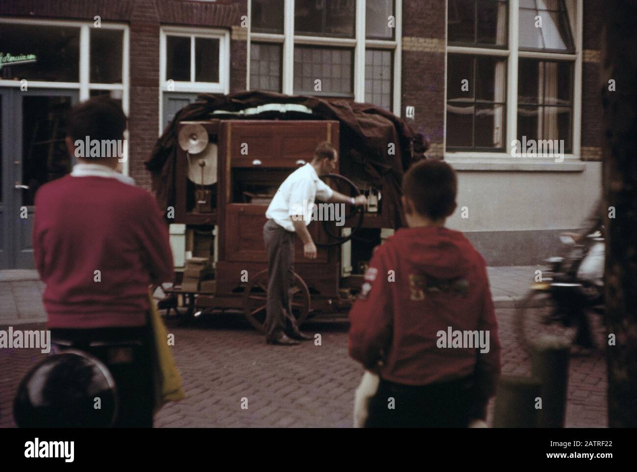 Vernacular photograph taken on a 35mm analog film transparency, believed to depict man in red shirt standing near brown wooden cabinet, likely an organ grinder or one man band show, 1970. Major topics/objects detected include Street. () Stock Photo