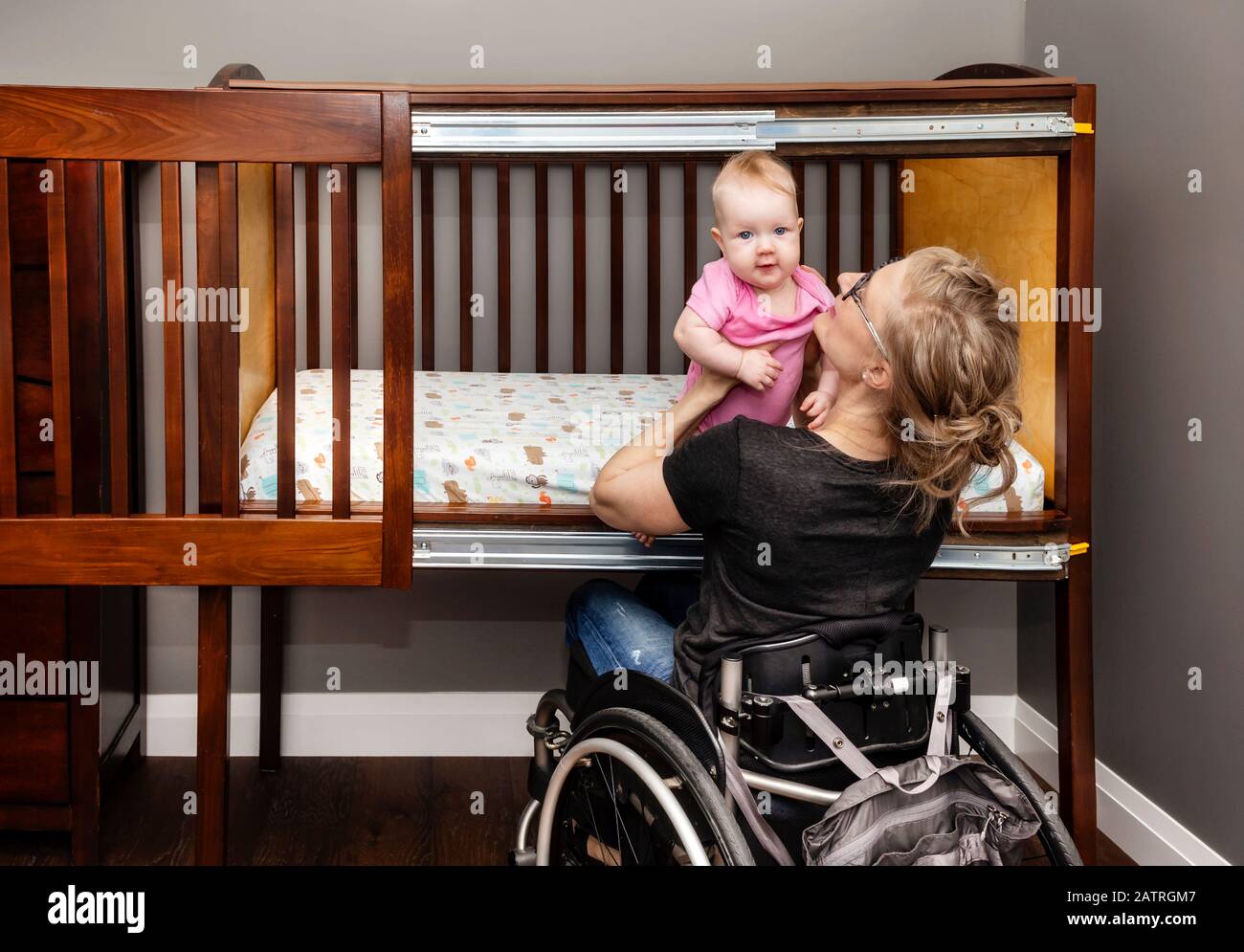 A paraplegic mother lifting a baby from a customized side-opening crib that allows her to put her baby down for a nap from her position in a wheelc... Stock Photo