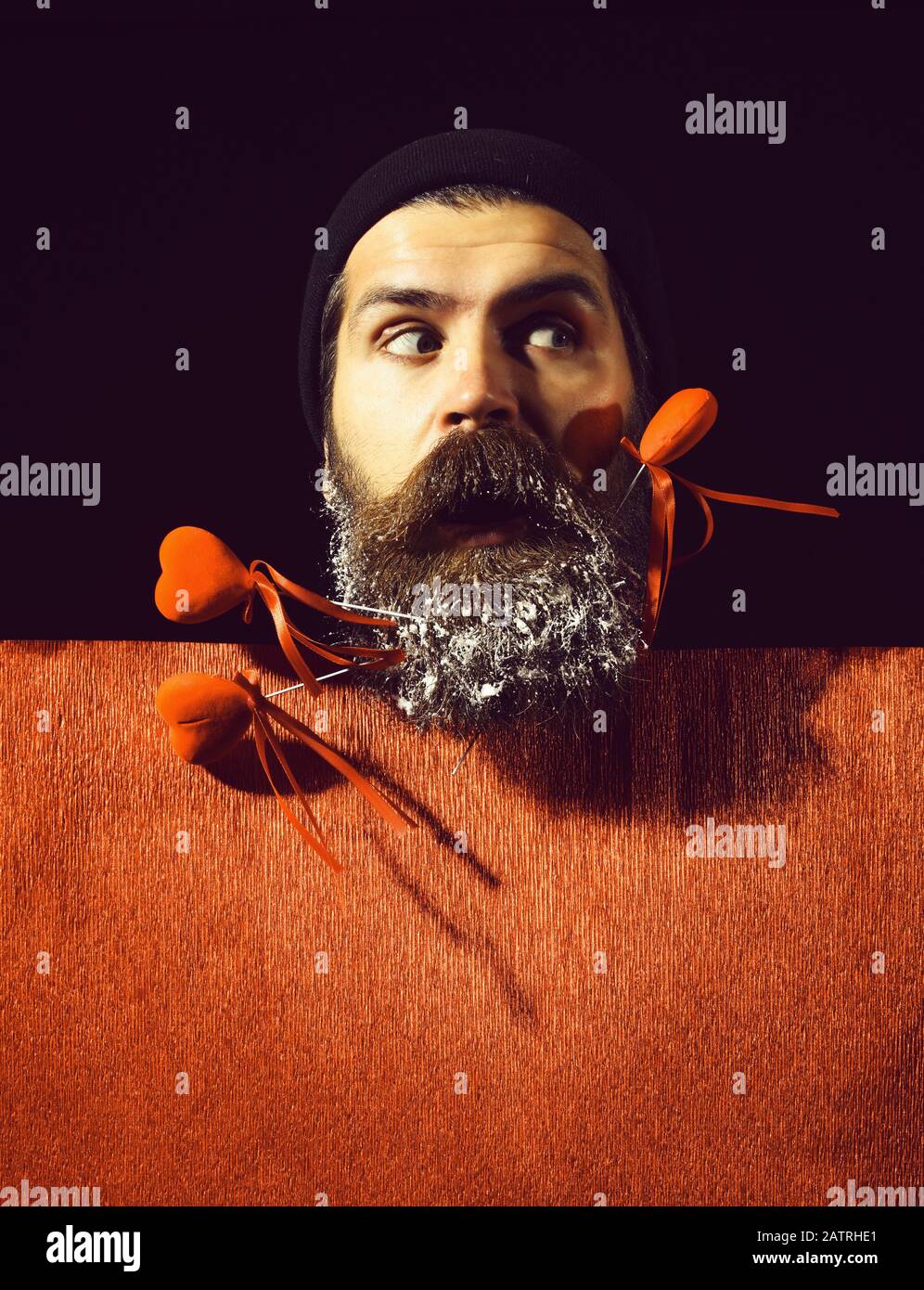 handsome bearded man or guy in winter hat with fashionable mustache on surprised face and snow in beard with decorative valentines hearts on sticks near red pape,r on black background, copy space Stock Photo