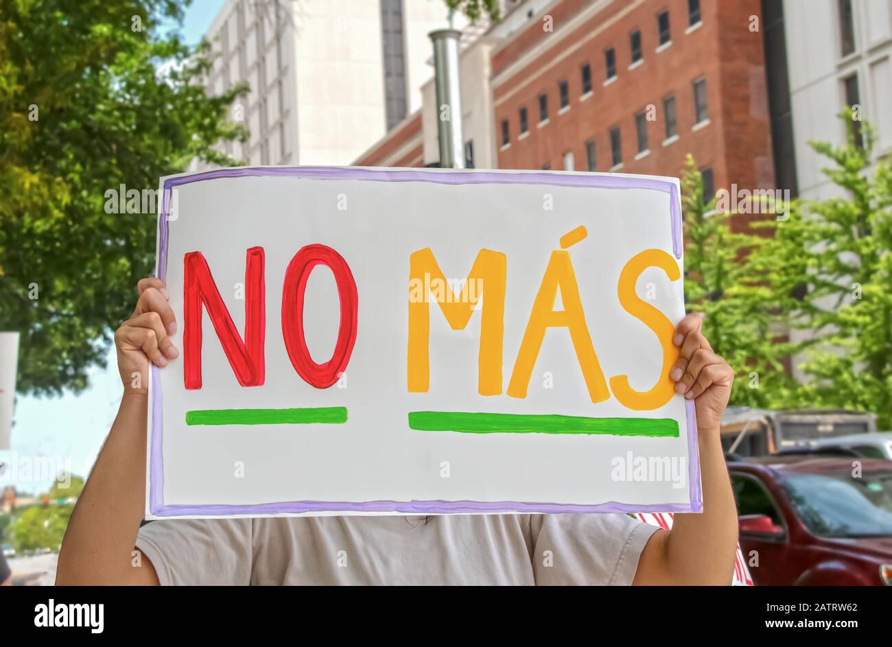 Woman holding up sign that says No Mas - Spanish for No More - in front of her face with tall buildings and cars in the background and another protest Stock Photo