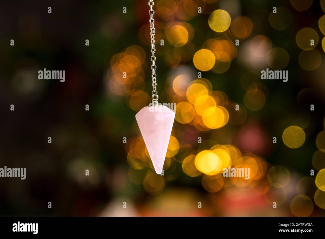 Pink color love rose quartz natural crystal pendulum hanging on silver chain indoors with round yellow bokeh lights on background. New years predictio Stock Photo