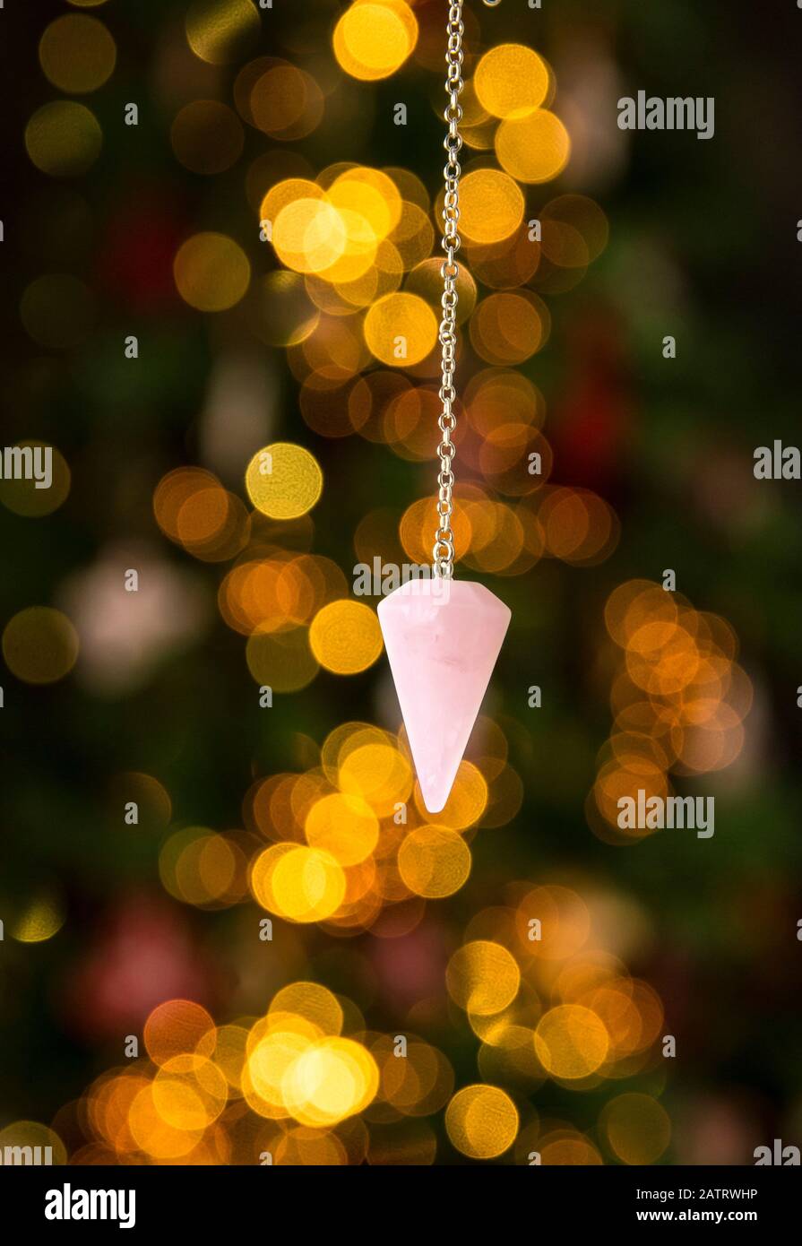 Pink color love rose quartz natural crystal pendulum hanging on silver chain indoors with round yellow bokeh lights on background. New years predictio Stock Photo