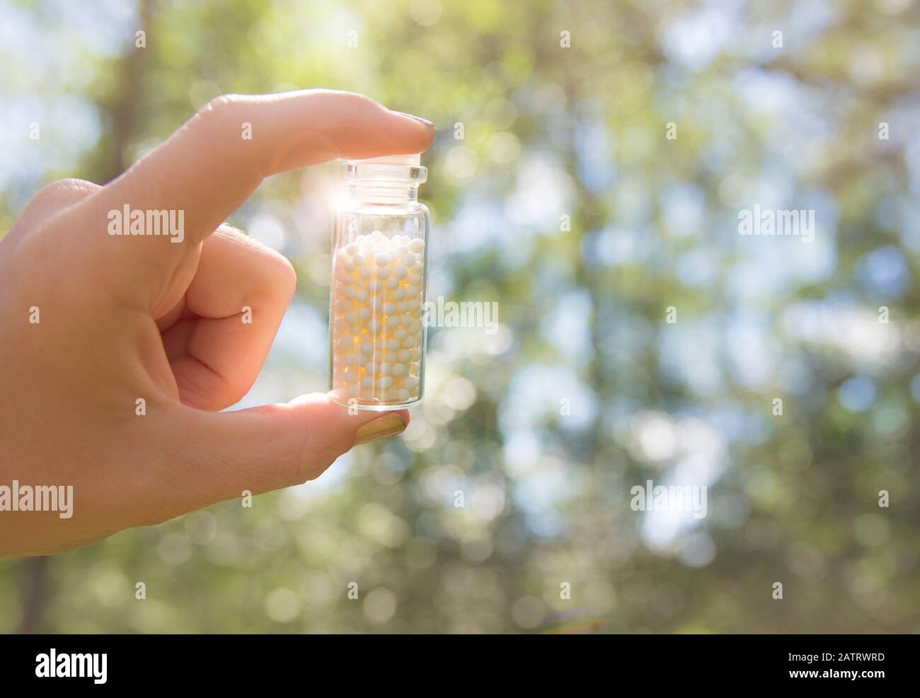 Selective focus on person hand holding glass jar full of small white round homeopathy pills against bokeh forest nature background in summer. Stock Photo