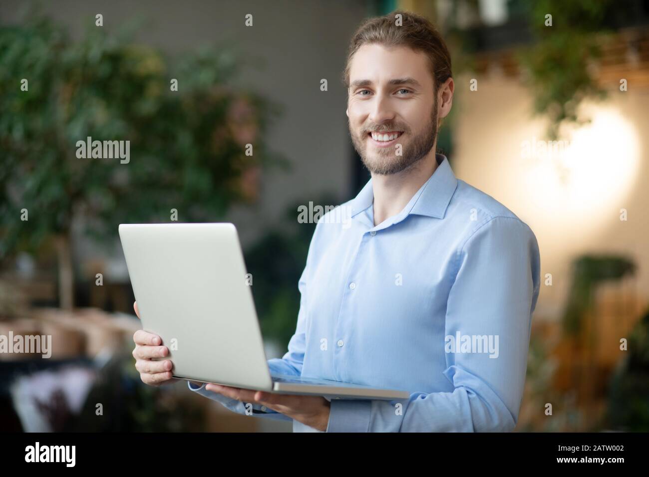 Good working day. Young smiling man in a light blue shirt holding a laptop in his hands, in a good mood. Stock Photo