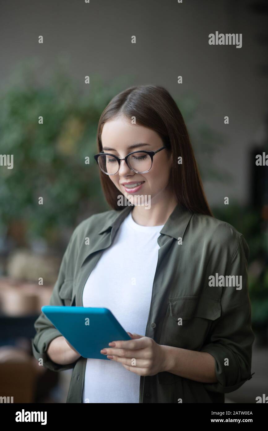 Tablet is always at hand. Successful smart girl in glasses with a blue tablet in her hands, looking at the screen, in a good mood. Stock Photo
