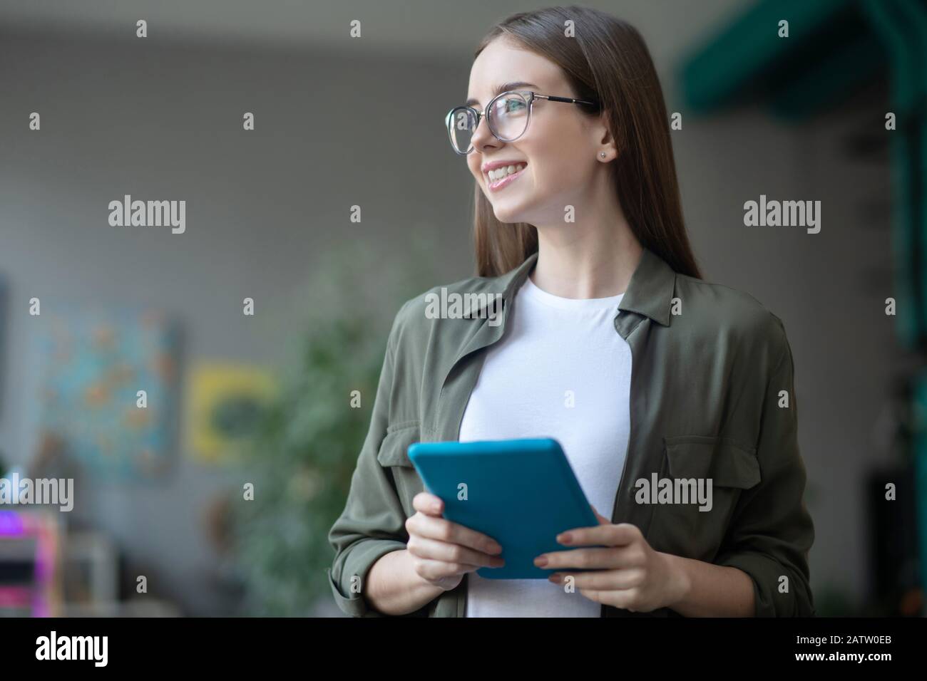 Great dreamy mood. Happy pretty girl with long dark hair in glasses with a tablet in her hands, looking to the side smiling and dreaming. Stock Photo