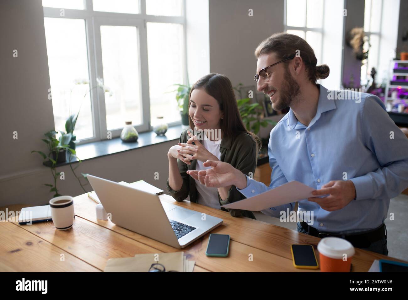 Business partners. Man with glasses and light blue shirt and girl with long dark hair working at a laptop in the office, smiling and joyful in good mo Stock Photo