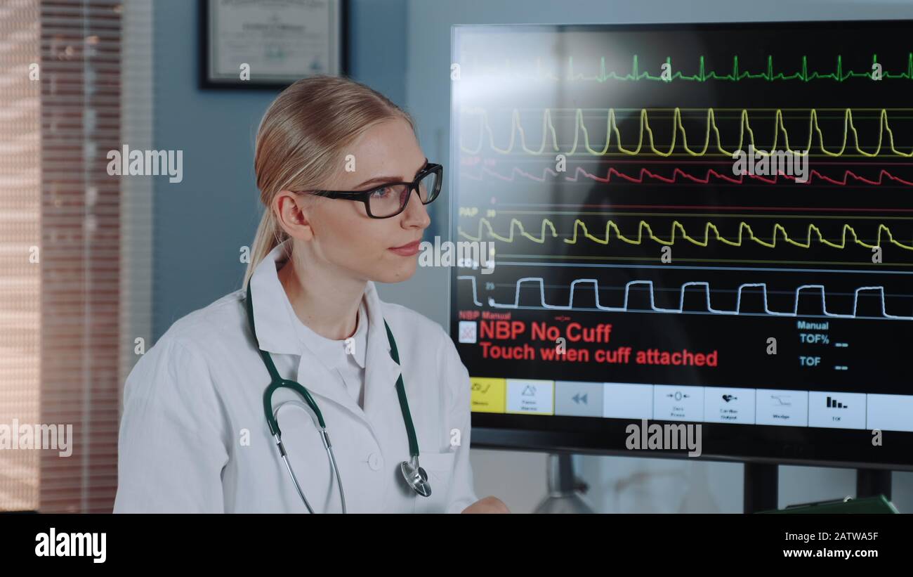 Close-up of beautiful female doctor in lab coat discussing EKG data with colleague showing on big display in modern sports lab. Stock Photo
