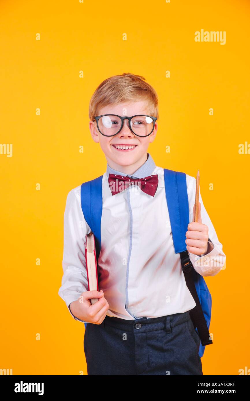 Happy cute clever boy in black glasses with blue school bag and book on yellow bakground. Stock Photo