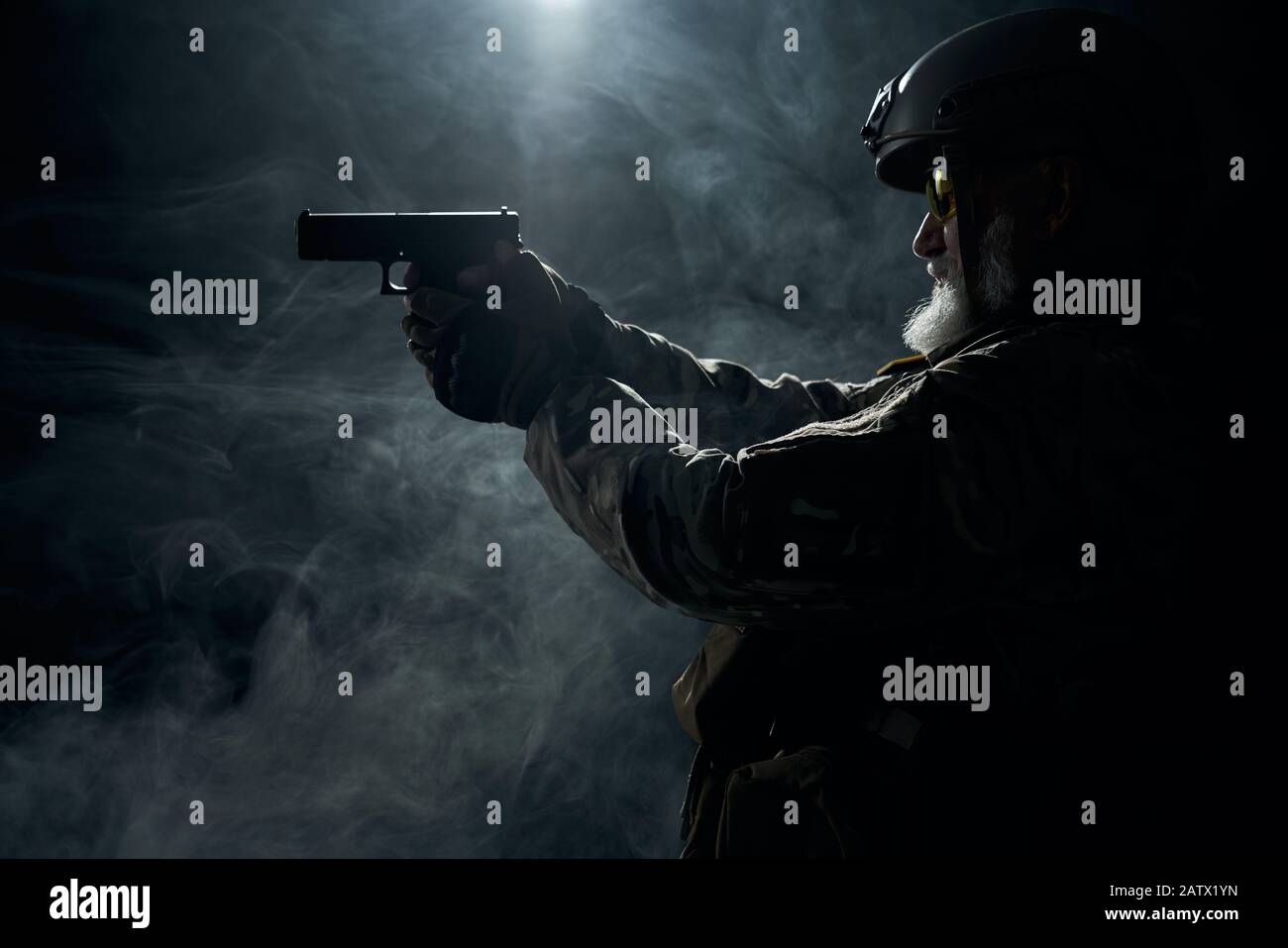 Side view of old military officer holding small gun and aiming. Portrait of bearded veteran in camouflage uniform and helmet shooting in smoky dark atmosphere. Concept of military. Stock Photo