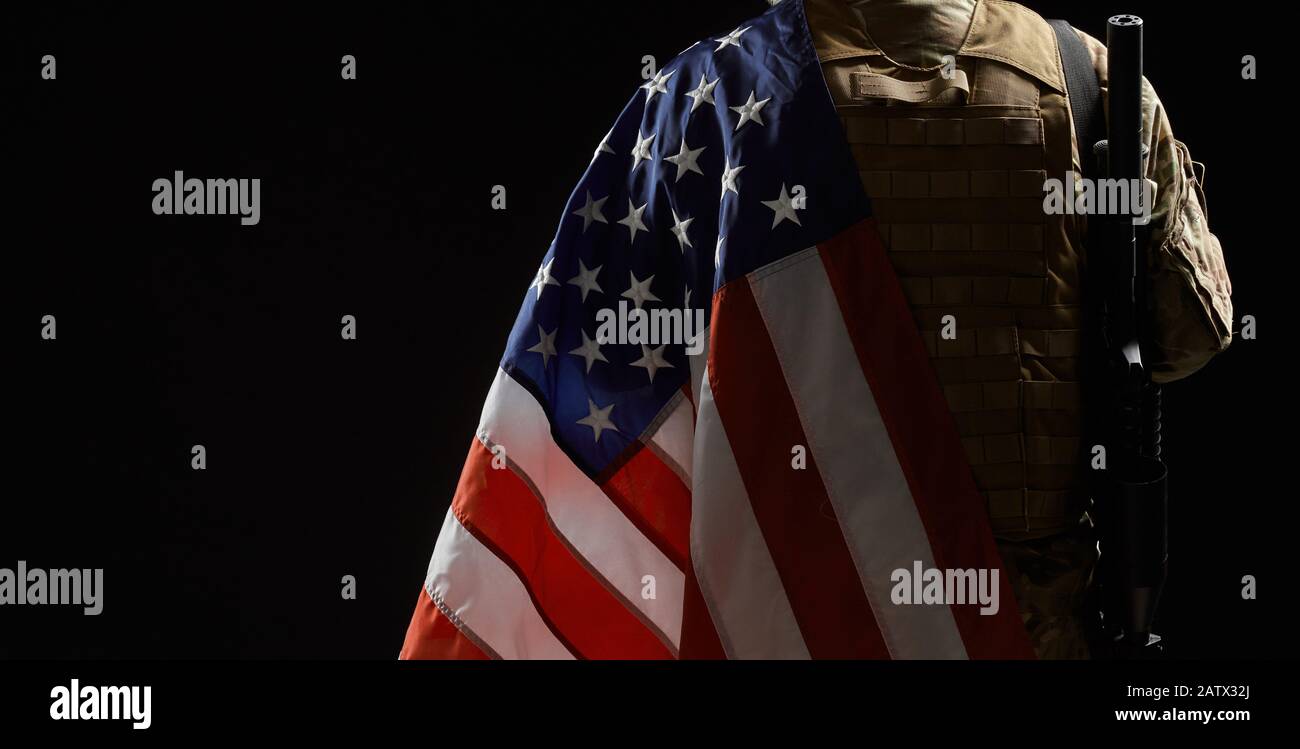 Back view of american military soldier holding flag and rifle on shoulder. Crop of incognito male officer in camouflage uniform posing in dark atmosphere. Concept of military, army. Stock Photo