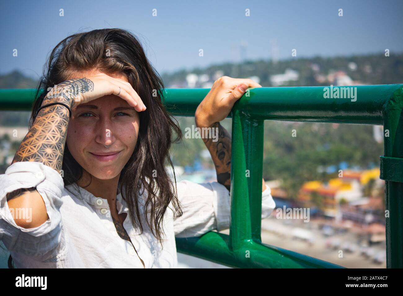 Beautiful girl with many tattoos on both arms shields her face from the strong sunlight whilst looking at the camera with a great view Stock Photo