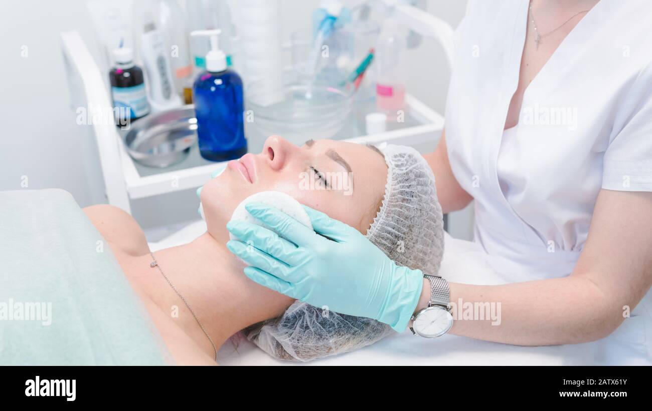 The female dermatologist cleaning face skin by cotton sponge to beautiful girl. Cosmetologist removing facial mask from female face. Concept of skin a Stock Photo