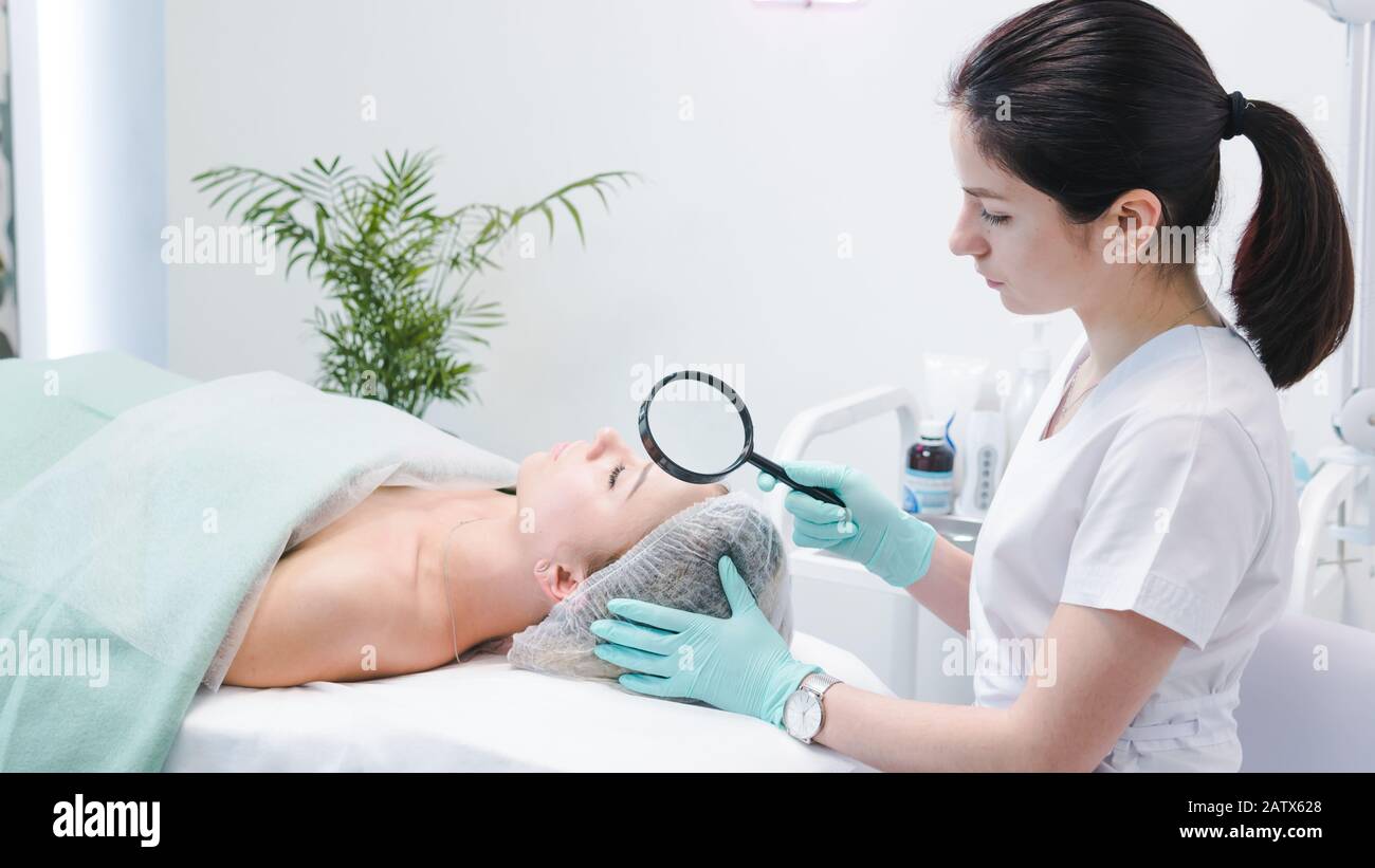 The female dermatologist examining face of young patient with magnifying glass in clinic or beauty salon. Concept of face cleanser, skin and health ca Stock Photo