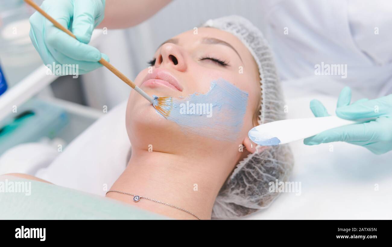 The female cosmetologist applying blue cosmetology mask to young woman face, close-up. Facial cosmetic treatment in beauty clinic. Concept of skin and Stock Photo