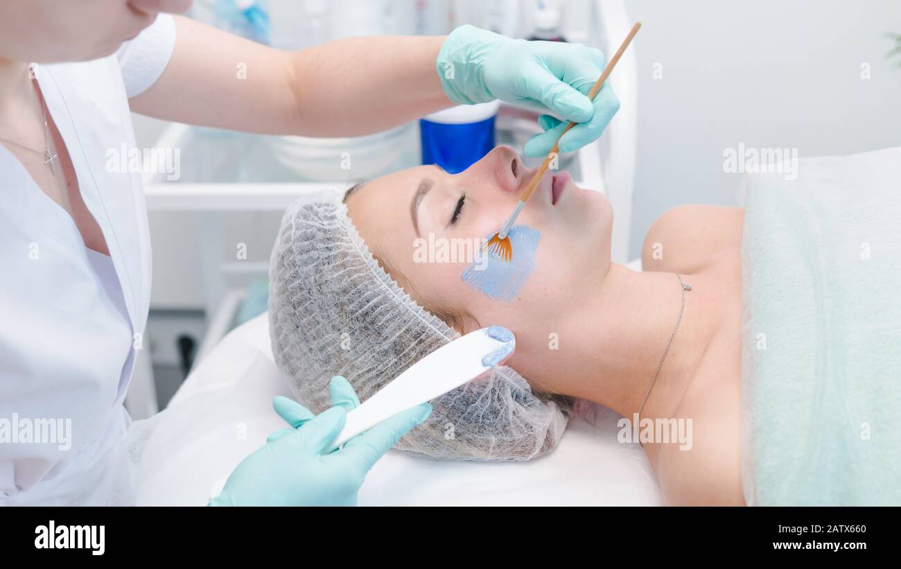 The female cosmetologist applying blue cosmetology mask to young woman face, close-up. Facial cosmetic treatment in beauty clinic. Concept of skin and Stock Photo