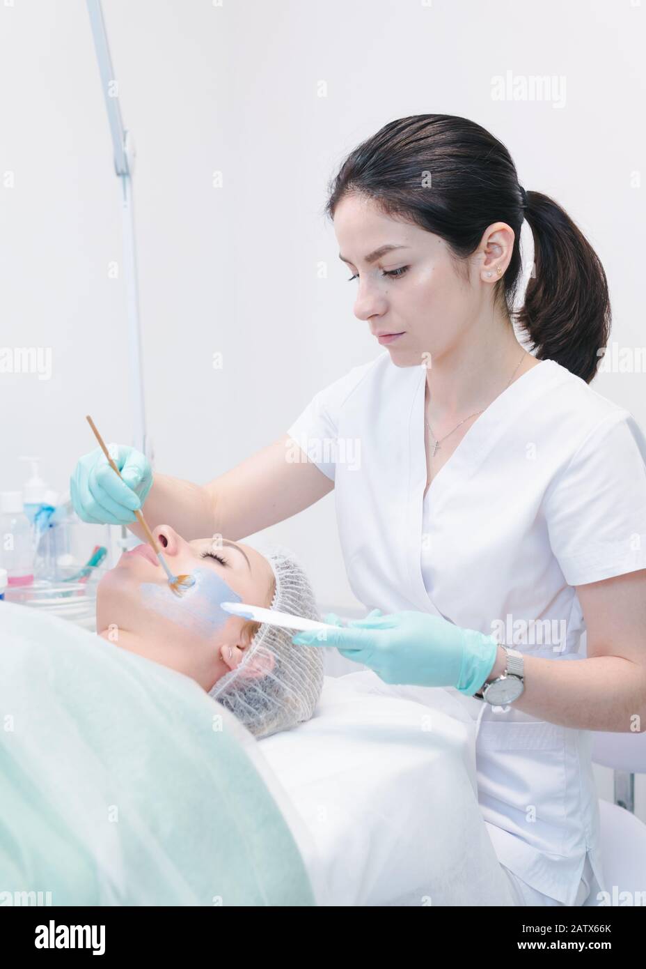 The female cosmetologist applying blue cosmetology mask to young woman face, close-up. Facial cosmetic treatment in beauty clinic. Concept of skin and Stock Photo