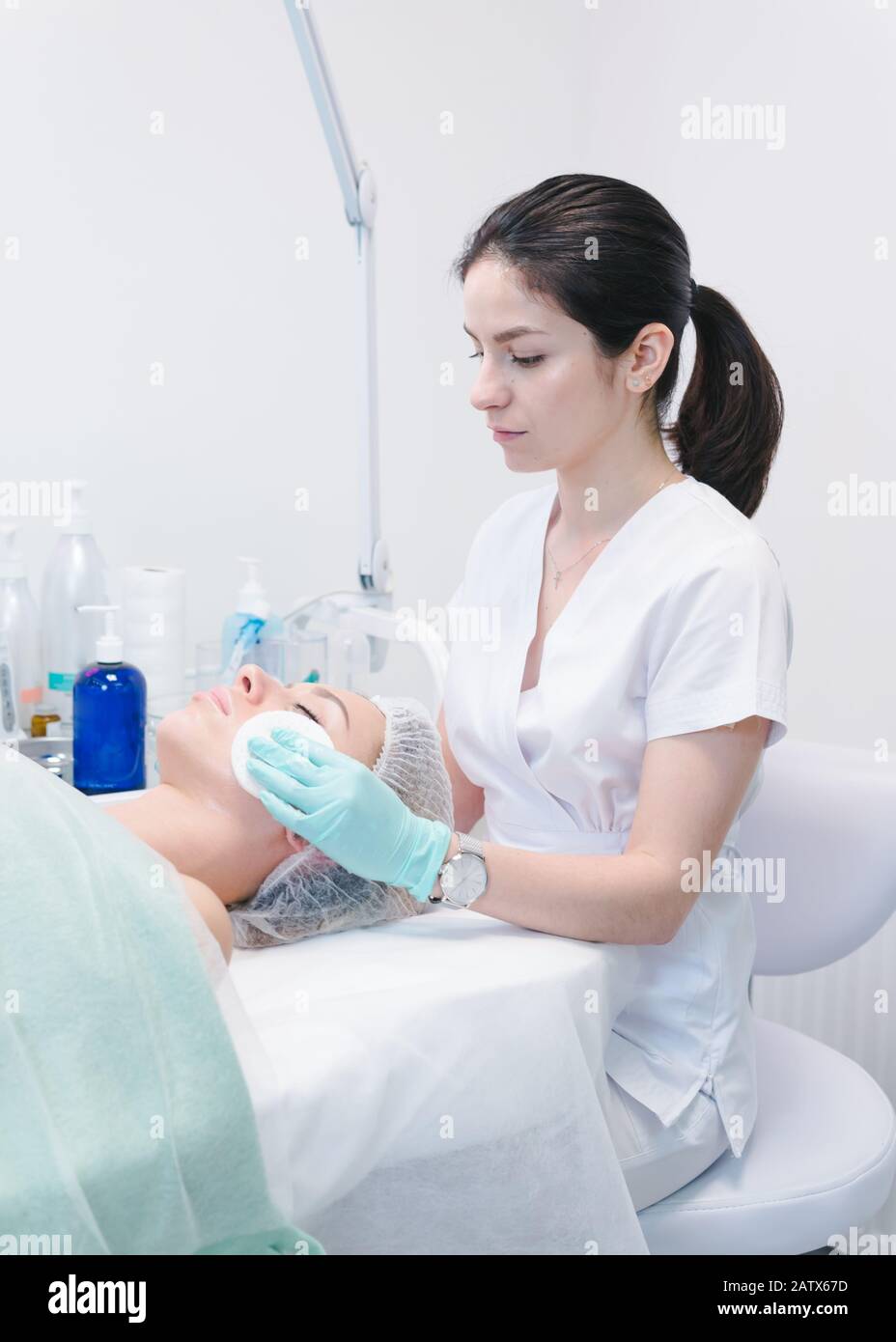 The female dermatologist cleaning face skin by cotton sponge to beautiful girl. Cosmetologist removing facial mask from female face. Concept of skin a Stock Photo