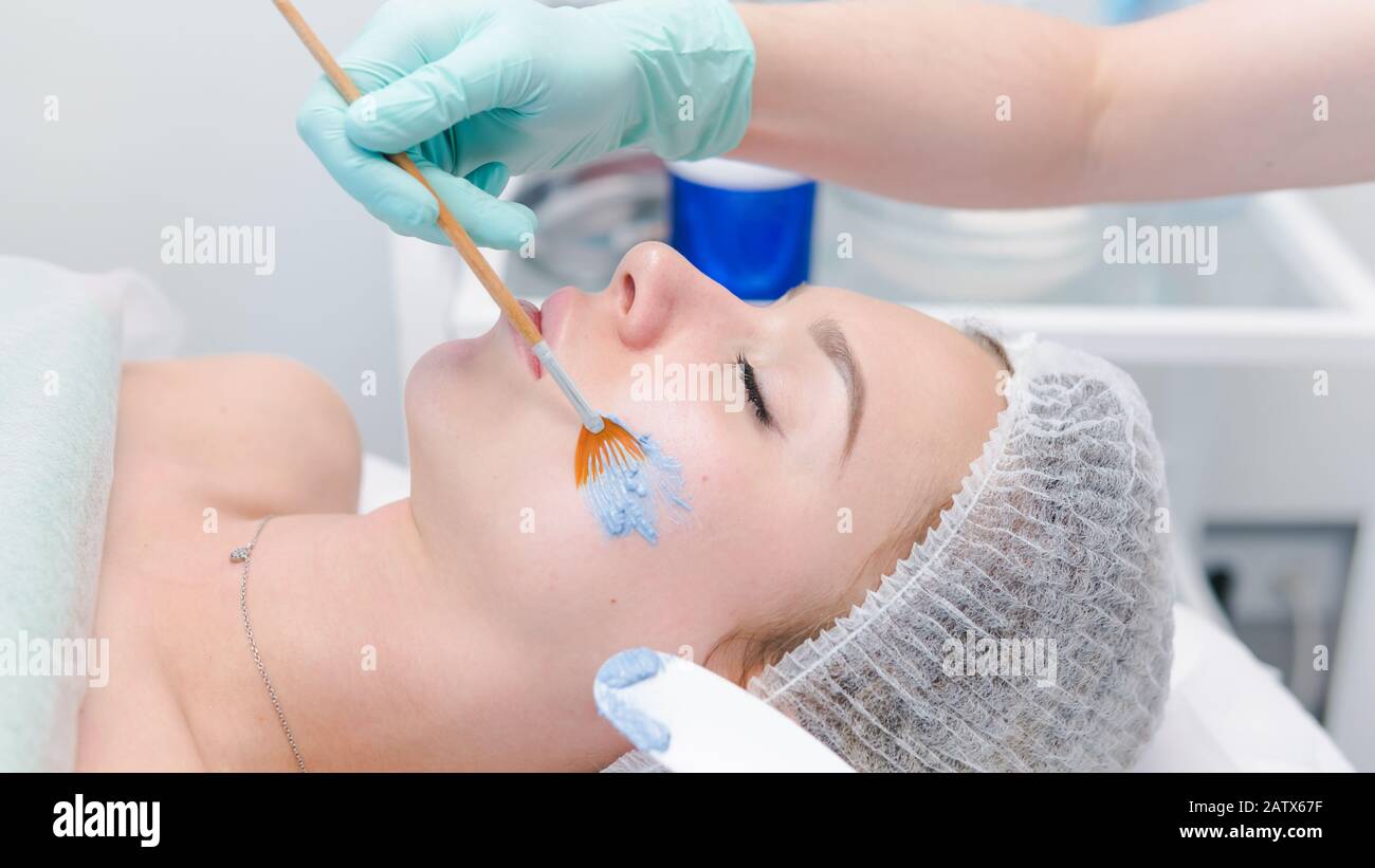 The female cosmetologist applying blue cosmetology mask to young woman face, close-up. Facial cosmetic treatment in beauty clinic. Concept of skin and Stock Photo