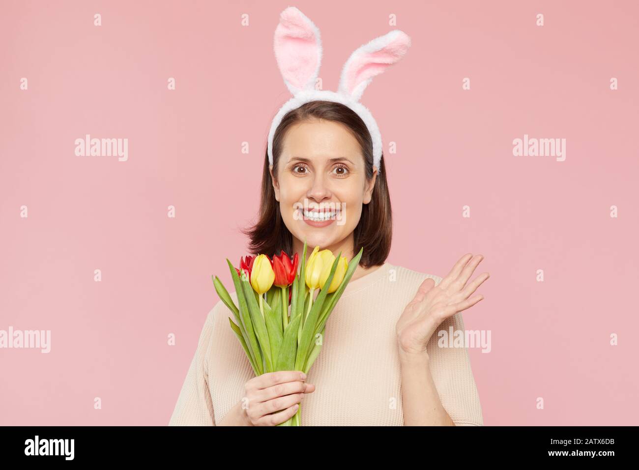 Portrait of happy young woman in rabbit ears holding bouquet of tulips and smiling at camera against the pink background Stock Photo