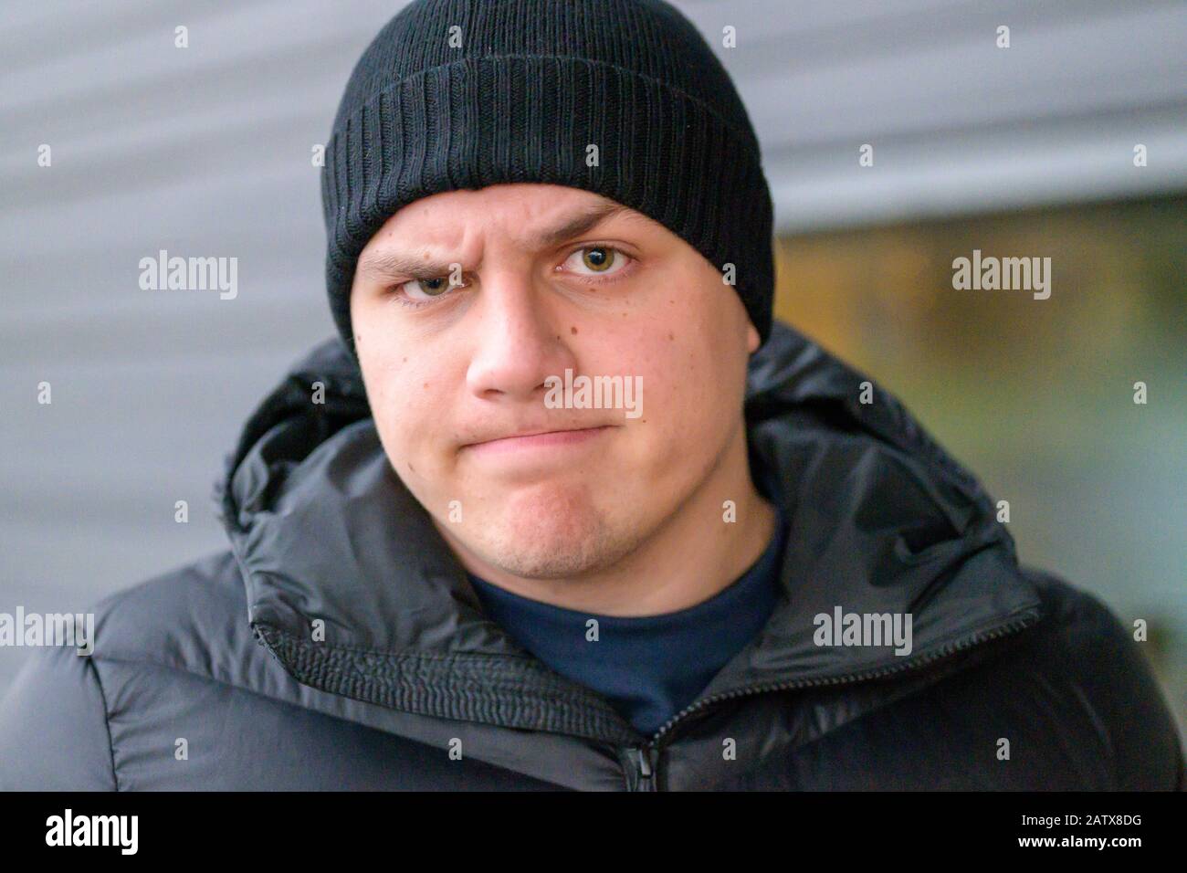 Determined sceptical young man wearing a beanie and warm jacket standing outdoors in winter frowning at the camera with raised eyebrow and a grimace Stock Photo