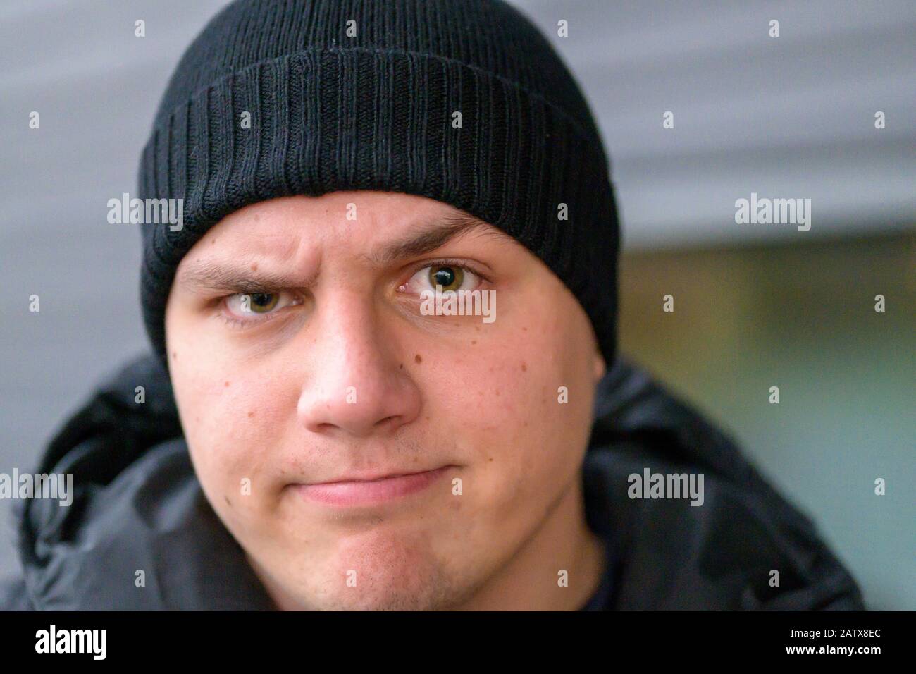 Determined sceptical young man wearing a beanie and warm jacket standing outdoors in winter frowning at the camera with raised eyebrow and a grimace Stock Photo