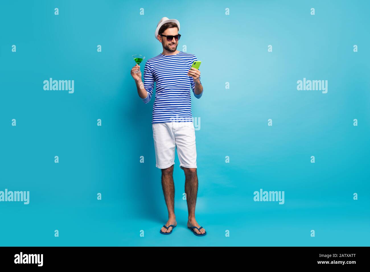 Full body photo of funky traveler guy browsing telephone drink green cocktail all inclusive resort wear sun specs striped sailor shirt cap shorts flip Stock Photo