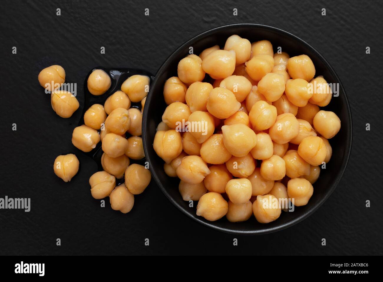 Cooked chick peas in a black ceramic bowl isolated on black painted wood. Spilled chick peas. Top view. Stock Photo