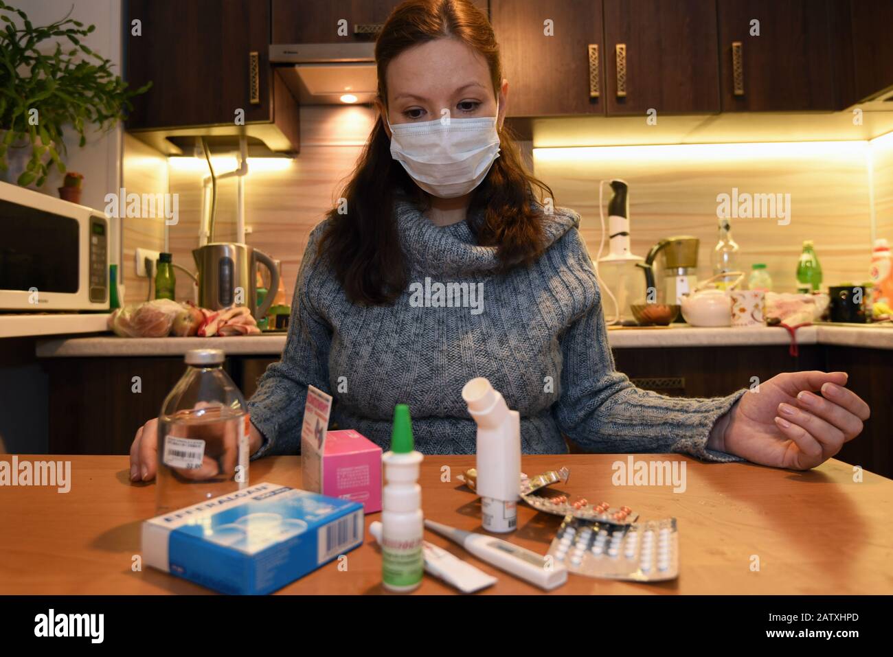 Woman in the mask being ill with a flu choses the medicaments in the kitchen. Stock Photo