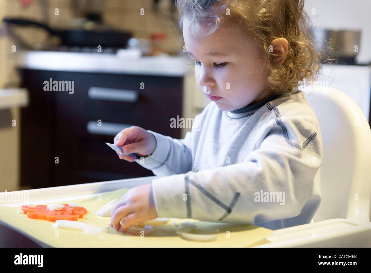 Kid cuts carrots and potatoes in kitchen. Cute two-year little girl helps the mother to cook. Independent little child learns to cook food. Beautiful Stock Photo