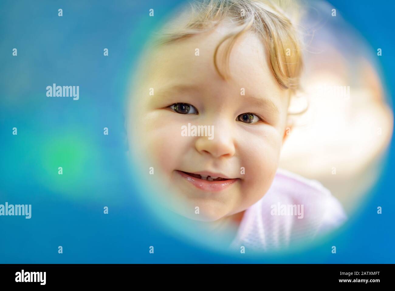 The one-year child plays on the playground. Cute baby girl looks through a round window to the camera. Stock Photo