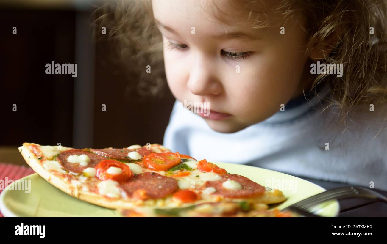 Child looks at pizza on table. Three years old kid is going to eat by himself. Little girl and food on plate. Feeding cute baby at home. Portrait of n Stock Photo
