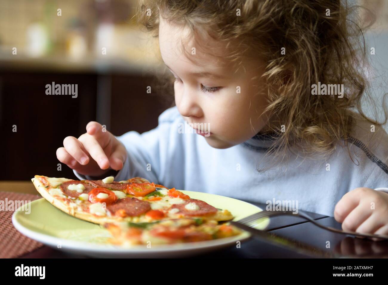 Child looks at pizza on table. Three years old kid is going to eat by himself. Little girl wants to take a piece of food from plate. Feeding cute baby Stock Photo