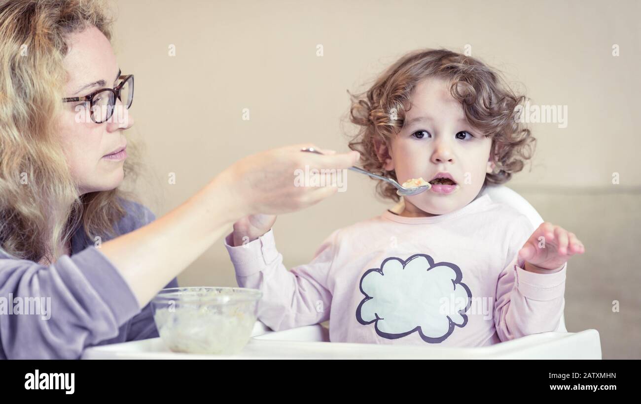 Baby girl during eating at home. Mother feeds her cute baby with a spoon. Adorable toddler sits on high chair and has problem with food. The two-year- Stock Photo
