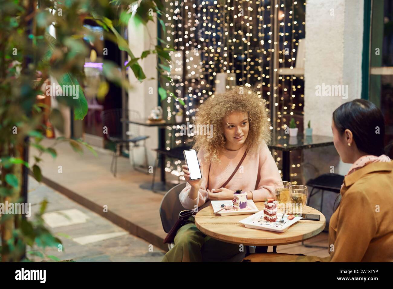 Young woman holding mobile phone and showing photos to her friend during their lunch in cafe Stock Photo