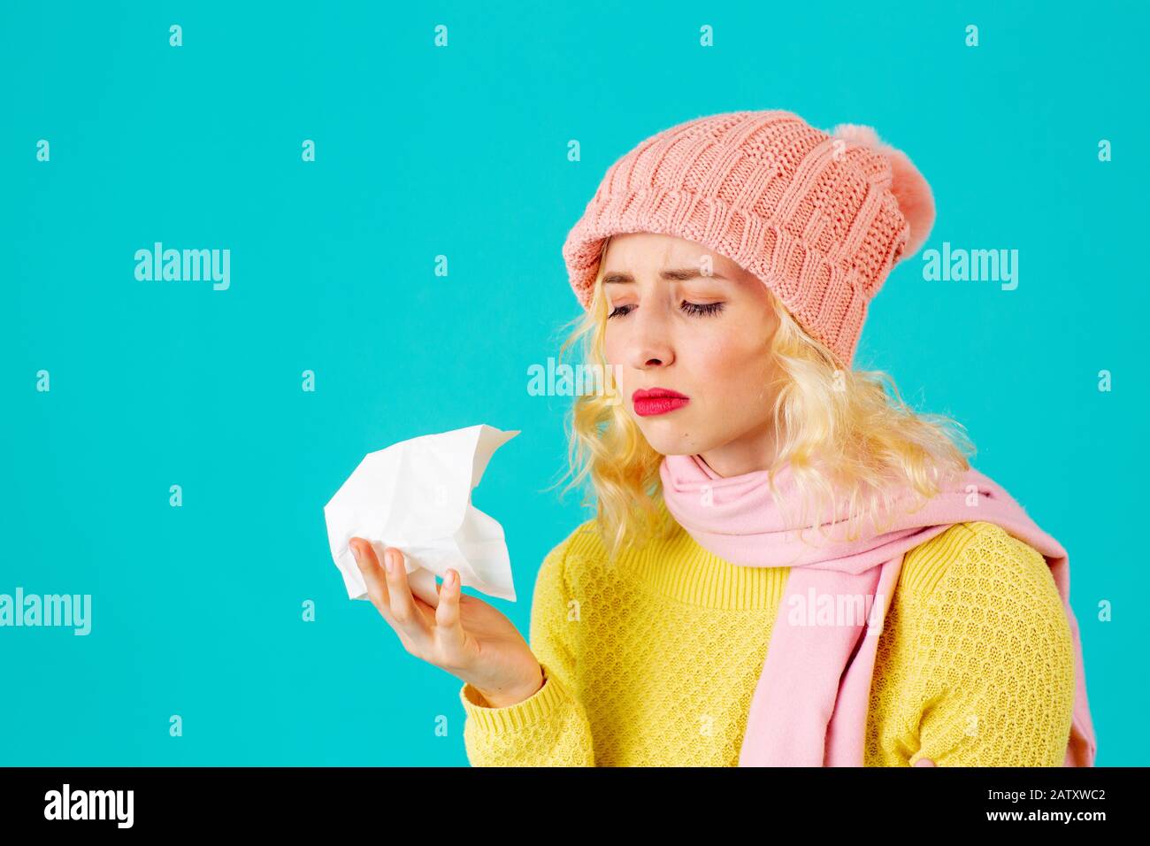 Cold and flu season- portrait of a woman in hat and scarf holding tissue isolated on blue Stock Photo