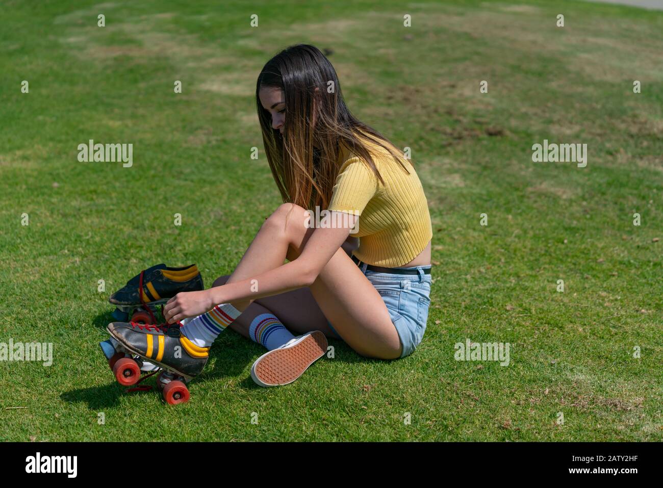 Pretty teenage girl in yellow top sits on grass putting retro style roller skates on getting ready to go skating. Stock Photo