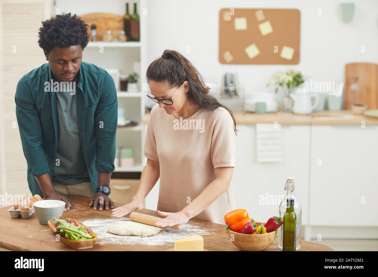 Young woman in eyeglasses standing and rolling dough with her boyfriend helping her they are in the kitchen at home Stock Photo