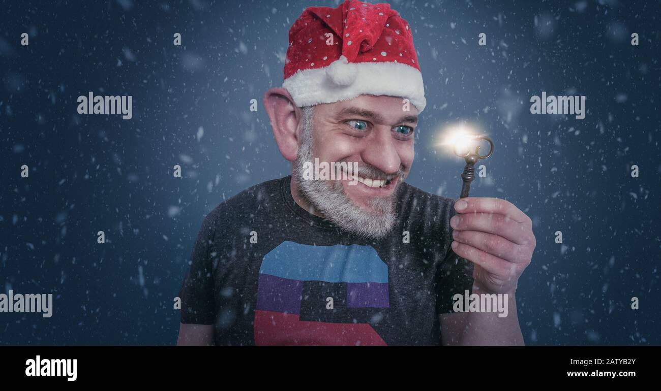 Bearded man in a Santa hat at Christmas holding a glowing key in his hand with a gleeful smile in close up with falling snow at twilight Stock Photo