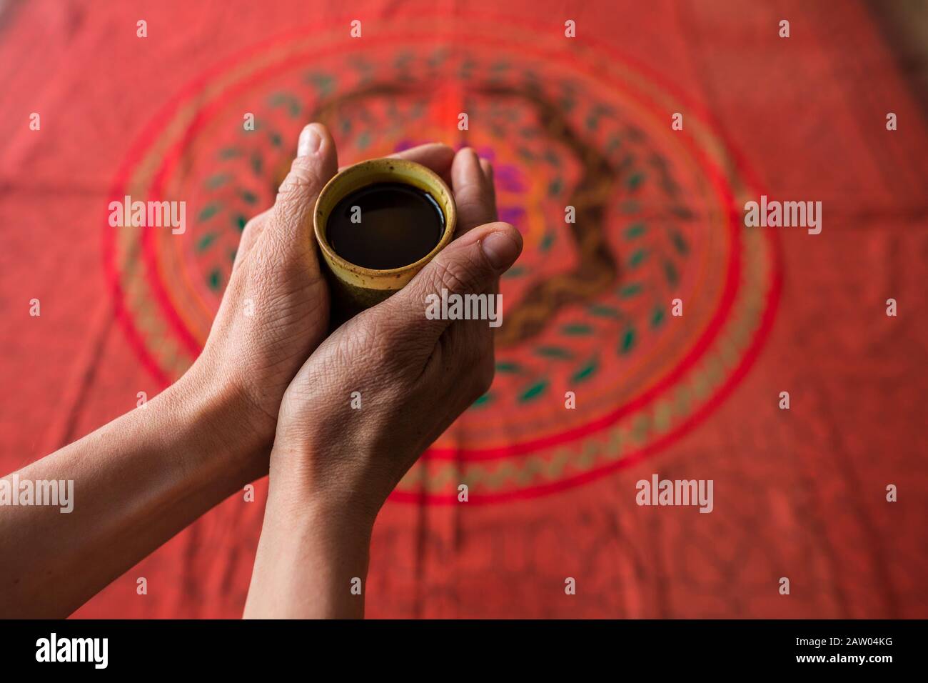 A man holds a cup of hallucinogenic Ayahuasca plant medicine tea in his outstretched hands over a Shipibo embroidered ceremonial tapestry. Stock Photo
