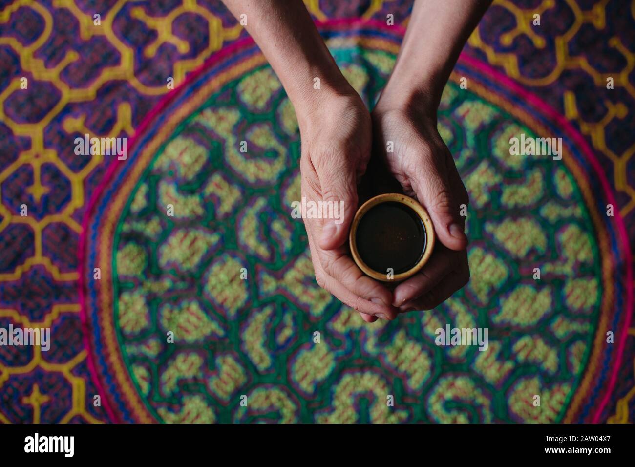 A man holds a cup of hallucinogenic Ayahuasca plant medicine tea in his outstretched hands over an embroidered Shipibo ceremony tapestry. Stock Photo