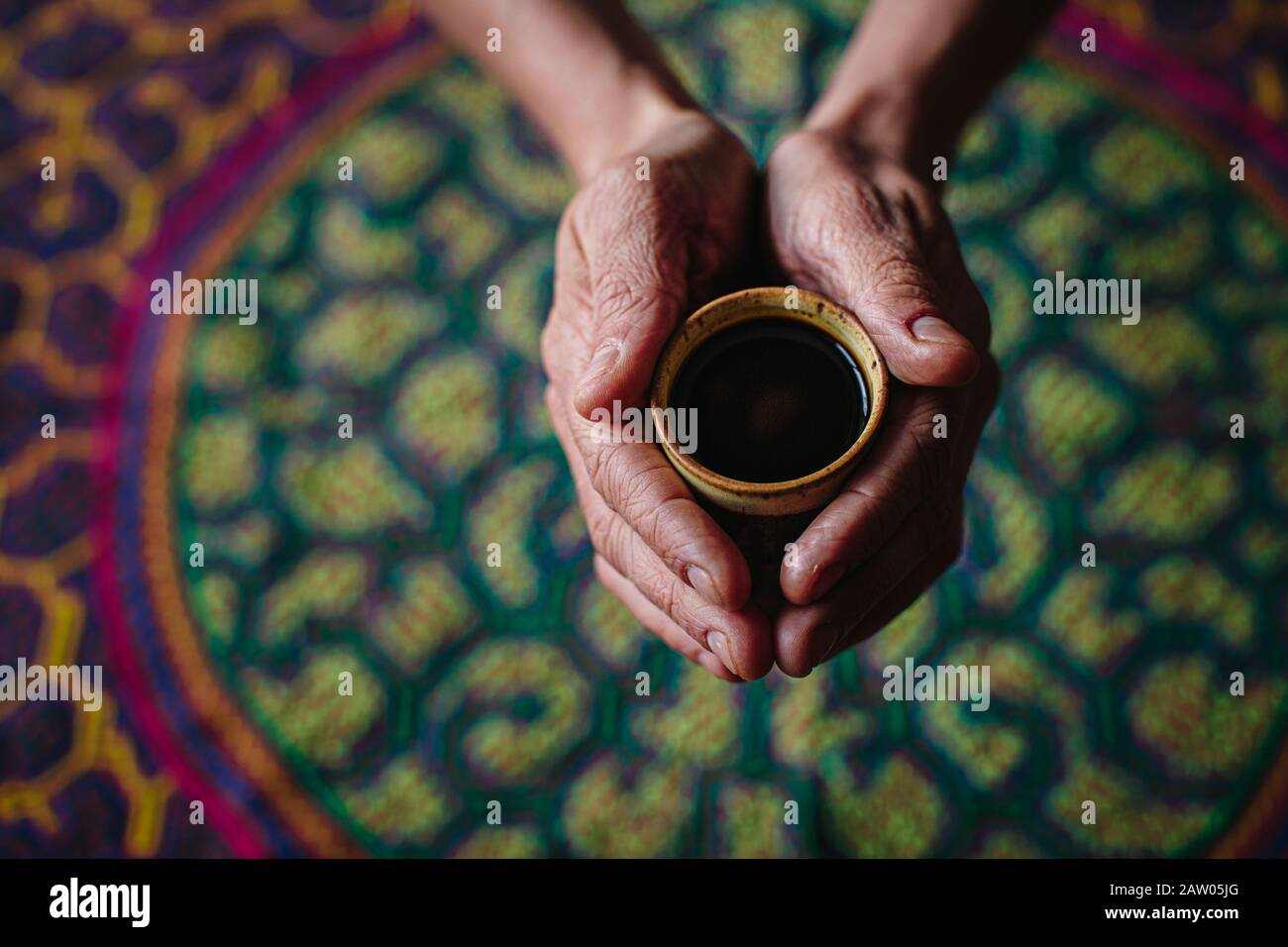 A man holds a cup of hallucinogenic Ayahuasca plant medicine tea in his outstretched hands over an embroidered Shipibo ceremony tapestry. Stock Photo