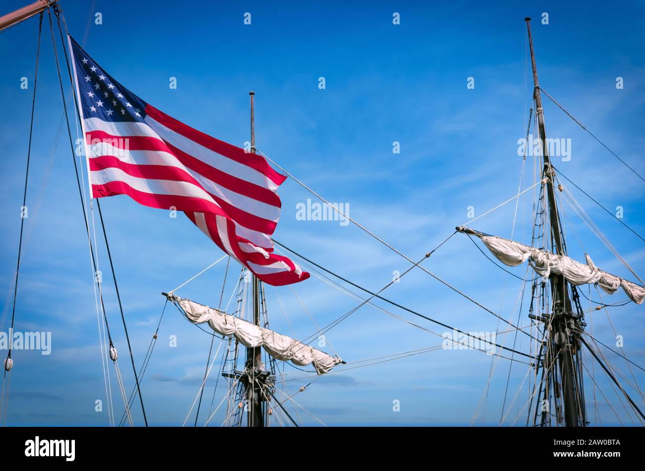 The American flag blows in the wind with ship masts and sails in the background. Stock Photo