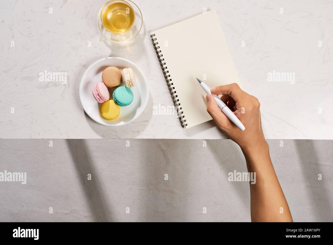 Man writing on notebook with cake macaron and tea mug on white background. Flatlay Stock Photo