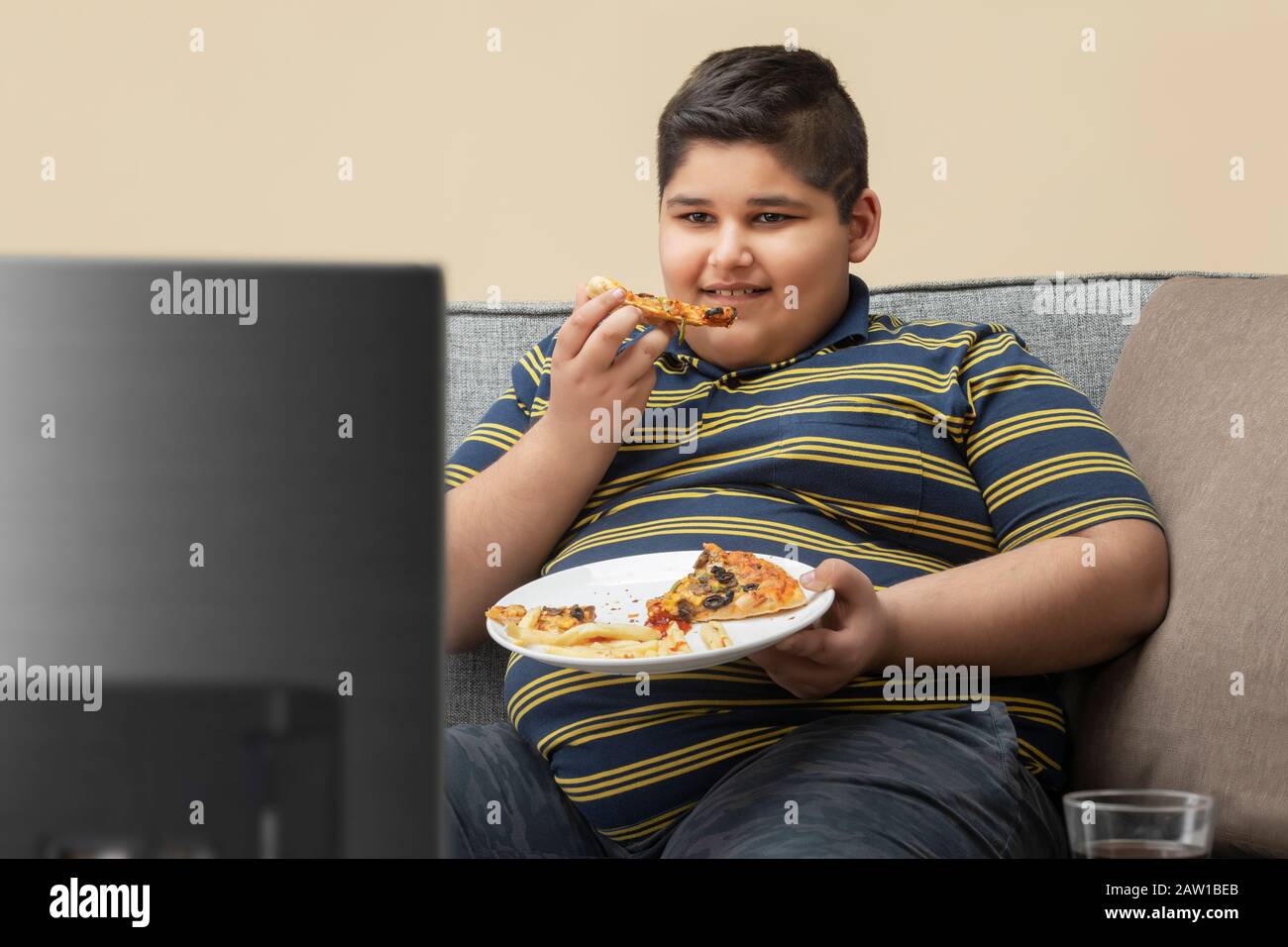 Young boy sitting on the couch at home and watching television while eating pizza. (Obesity) Stock Photo