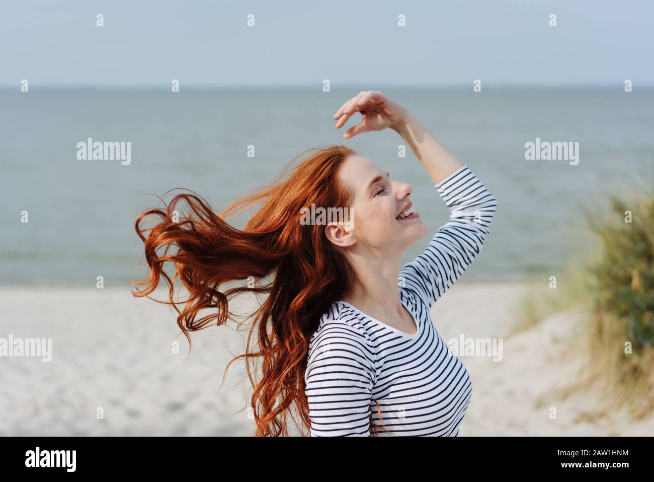 Carefree young woman tossing her long red hair with a happy smile in late evening sun on a winter beach with ocean backdrop Stock Photo